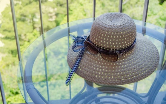 Summer straw hat and sunglasses close-up on a glass table on the balcony.