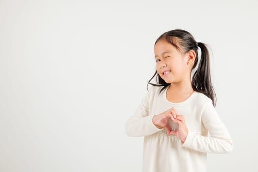 A smiling Asian young kid girl shows confidence, creating a heart shape with fingers and hands on white background. Portrait of kindergarten child sending love for Valentine's Day