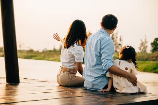 Asian family of three a girl child are sitting on a wooden dock by a body of water. Happy parent father mother and daughter kid they are all smiling. Concept of family warmth and happiness
