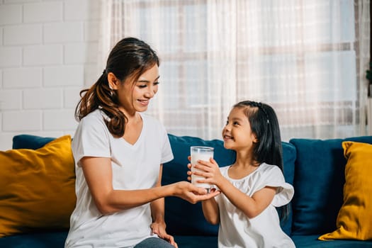 In their modern home an Asian mother lovingly offers her daughter a glass of milk on the sofa. This image reflects their carefree happiness togetherness and calcium-rich diet.