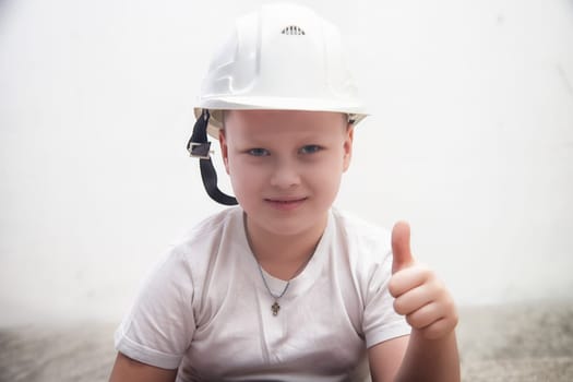 A child wearing a white construction helmet smiles and gives a thumbs up in a well-lit room, conveying excitement and positivity.