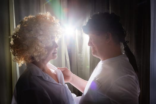 A middle-aged couple showcases their love while posing in vintage wigs during a specialized photo shoot in their apartment.