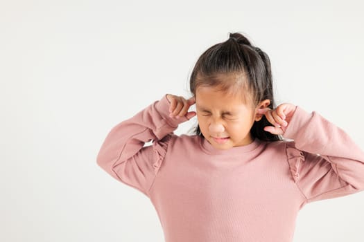 Asian young kid girl expression of pain and frustration is evident as she covers ears and closes eyes with fingers to escape the loud noise in studio shot isolated white background, kindergarten child