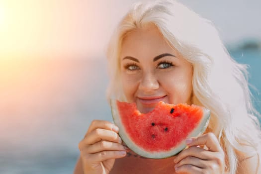 Smiling blonde woman with long hair holds a large slice of red watermelon to her face near the ocean.
