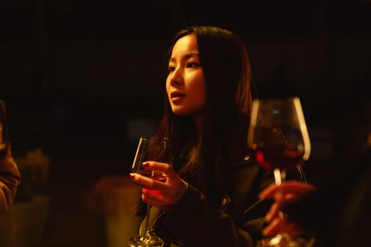 In a cozy venue with soft, warm lighting, a young woman holds a glass of wine and engages in conversation with others nearby, creating a relaxed atmosphere.