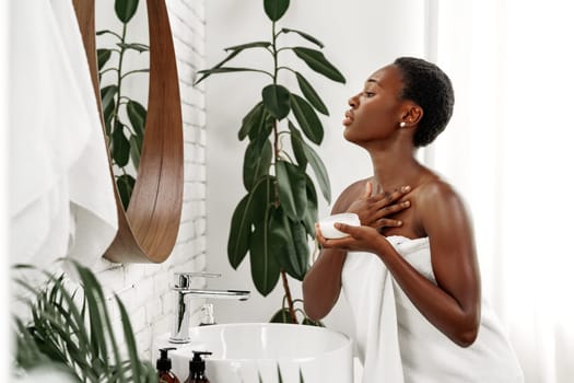 A woman wraps her body in a towel while applying cream in front of a round mirror. Sunlight streams through the window, illuminating her skincare routine and the plants nearby.