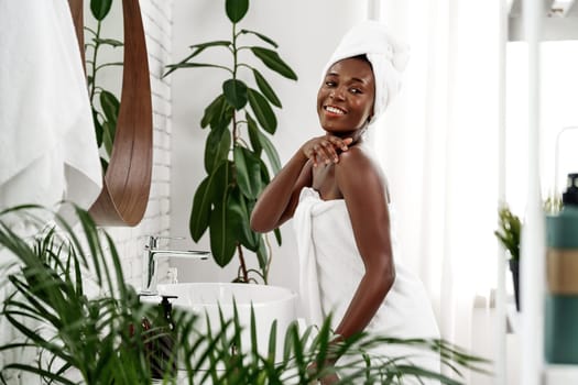 A joyful woman with a towel around her body smiles while applying lotion in a stylish bathroom filled with plants and bright sunshine. The atmosphere exudes tranquility and wellness.