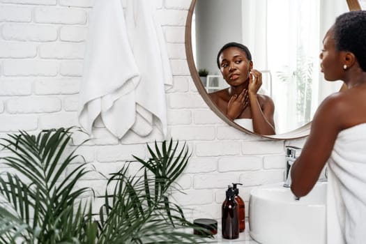 Young woman with short hair applies skincare products while looking in a round mirror. The bright bathroom features white brick walls and lush indoor plants, creating a refreshing atmosphere.