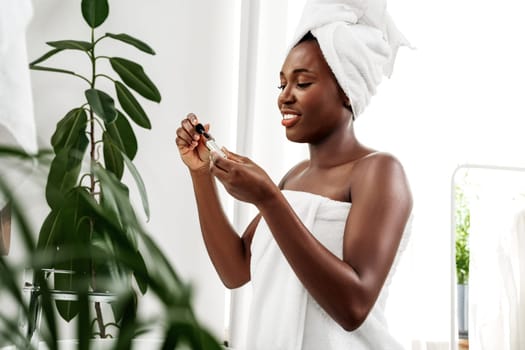 A woman wrapped in a towel smiles as she applies a skincare serum in a well-lit bathroom. Green plants add a natural touch to her relaxing self-care moment.