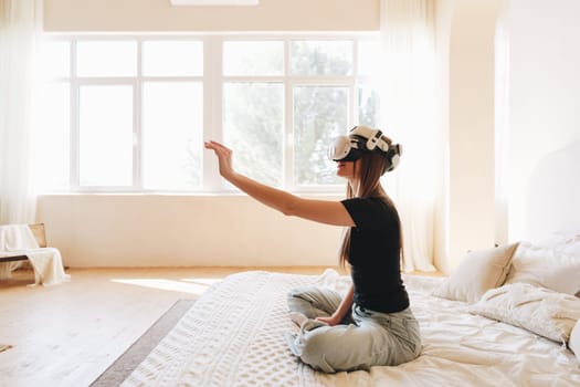 A woman sits cross-legged on a bed in a bright room, wearing a virtual reality headset and reaching out with her hand, exploring immersive technology.