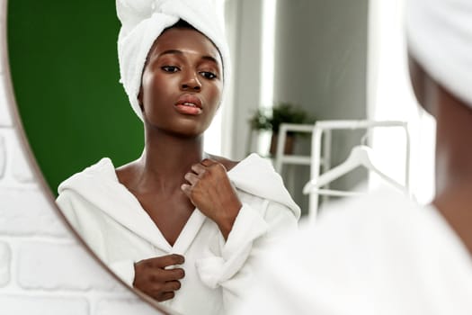 A young woman stands in front of a stylish mirror, wearing a white bathrobe and towel. She adjusts her head towel while gazing thoughtfully, surrounded by a clean, bright bathroom.