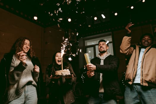 Group of friends gather indoors for a birthday celebration, holding a cake adorned with sparkler candles. They display excitement while enjoying the festive atmosphere, surrounded by decorations.