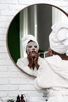 In a stylish bathroom, a woman in a white robe and towel observes herself in the round mirror while applying a facial mask, enjoying her self-care routine.