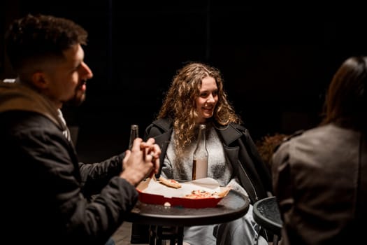 A group of friends relaxes at a small round table, laughing and sharing pizza under dim lighting on a chilly winter night in the city.