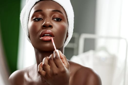 In a well-lit bathroom, a woman with a towel on her head applies lip gloss to her lips while looking into a mirror. Natural light enhances her glowing complexion.