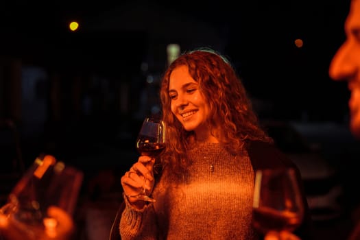 Friends gather on a cool evening, mingling and enjoying glasses of wine. Warm lights cast a cozy glow, enhancing the cheerful atmosphere of their outdoor get-together.