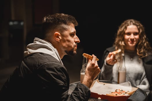 Two friends are seated at an outdoor table in a dimly lit cafe. They are enjoying late-night snacks, with one person holding a slice of food, sharing a light-hearted moment.