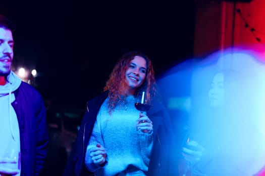 A group of friends shares a joyful moment during an outdoor event at night. Colorful lights illuminate the setting, enhancing the festive atmosphere while they hold drinks.