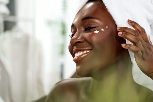 A happy woman with a towel on her head smiles while applying a skincare cream on her cheek in a bright bathroom. Natural light enhances the fresh atmosphere of her morning routine.