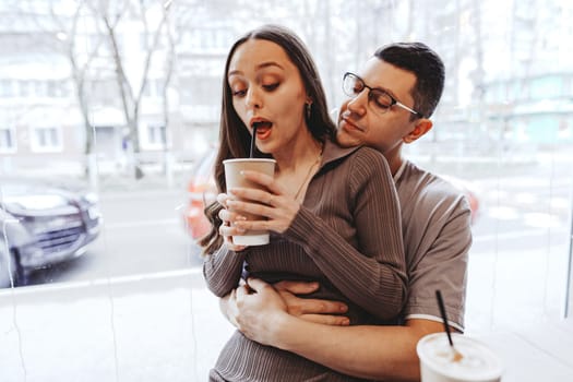 Inside a warm cafe, a couple shares drinks, with one partner playfully reacting to a sip. They sit closely, surrounded by a lively city backdrop on a chilly day, enjoying their time together.