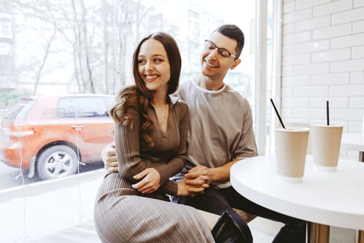 A couple shares a joyful moment in a bright cafe, smiling and looking at each other while seated at a small table. Two cups sit nearby, adding to the warm atmosphere.