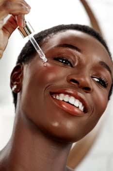 A woman with short hair smiles while applying a serum to her cheek using a dropper. The modern bathroom features natural lighting, enhancing her radiant skin.