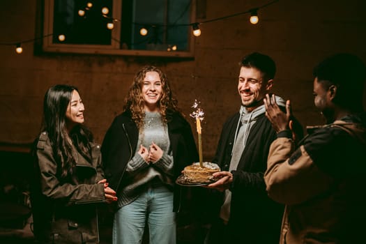 Friends gather outdoors to celebrate a birthday during the evening. The atmosphere is lively and festive, featuring a cake with a sparkler while they enjoy each other's company.