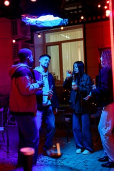 Four friends share laughter and drinks during a vibrant evening gathering, surrounded by colorful lights in a cozy outdoor space. The atmosphere is festive and joyful.