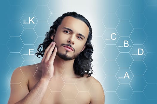 A man with curly hair touches his face while surrounded by a blue background representing vitamins A, C, D, E, and K. He appears confident and engaged in skincare knowledge.