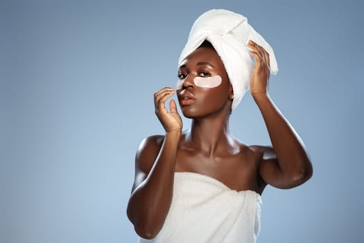 A young woman stands in a serene indoor space, draped in a towel, applying a skincare treatment under her eyes. She exhibits a calm and focused expression, showcasing her self-care routine.