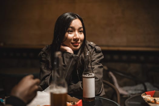 In a cozy atmosphere, a young woman rests her chin on her hand, deep in thought while surrounded by friends and drinks. The soft lighting creates an intimate vibe perfect for conversation.