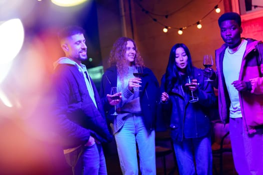 Four friends gather outdoors, holding wine glasses and smiling under vibrant lights. The cozy atmosphere fosters laughter and conversation on a lively night.