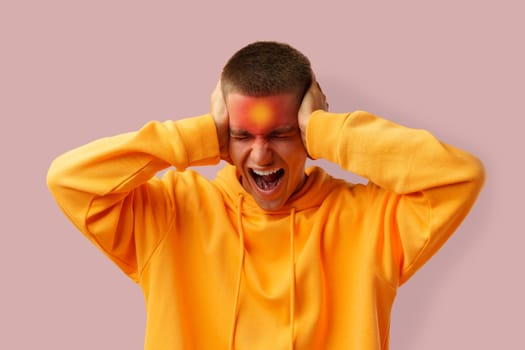 A young man is visibly stressed, his face contorted in frustration as he covers his ears with his hands. He wears a bright orange hoodie, set against a soft pink background.