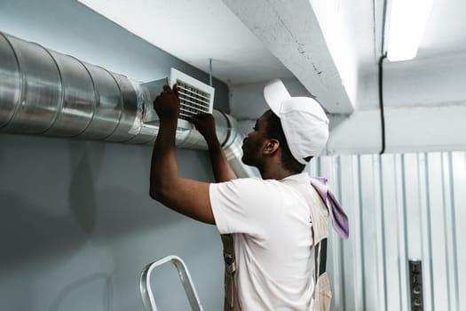 A technician adjusts an air vent while standing on a ladder in a commercial space. The setting features metal ductwork and ambient lighting. Attention to detail is evident in the careful work.
