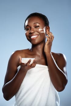 Happy woman with short hair enjoys applying skincare on her face while holding a small bowl of cream. Bright background enhances her radiant smile and glowing skin.