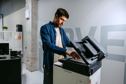 A man in a blue checkered shirt operates a multifunction printer in a contemporary office. He focuses on the task, showcasing a busy work environment.