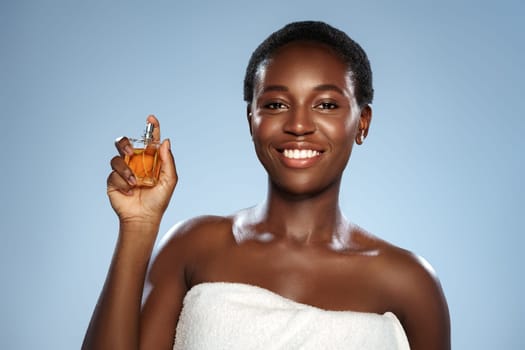 A joyful woman with short hair holds a small perfume bottle in one hand while wearing a towel. The soft blue backdrop enhances her radiant smile and the fragrance warm color.