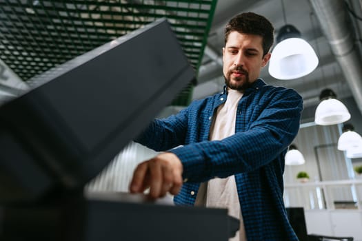 A young man in a blue shirt interacts with a sleek office printer, attentively managing the printing process in a contemporary workspace filled with soft lighting.