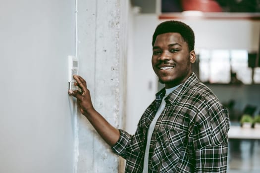 A young man with a cheerful expression is standing in a contemporary office, adjusting the thermostat on the wall. Sunlight fills the space, enhancing the positive atmosphere.