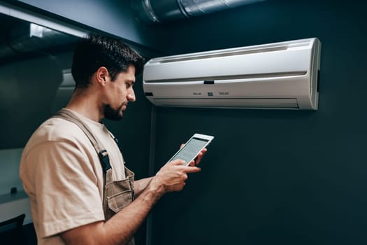 A technician stands close to an air conditioning unit, focused on checking information on his tablet. The setting is modern, with sleek walls and efficient lighting.