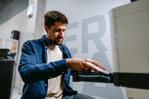 A focused man wearing a checkered shirt operates a printer in a contemporary office setting. He adjusts the machine with care, ensuring optimal functionality.