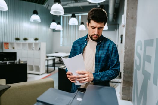 A man stands by a printer in a contemporary office, focusing on the papers he just printed. The workspace features modern design elements and ample lighting.