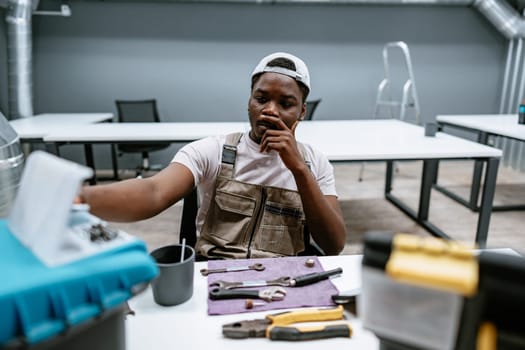 In a contemporary workshop, a young man wearing overalls thinks deeply while examining tools on a workspace with various equipment scattered around.