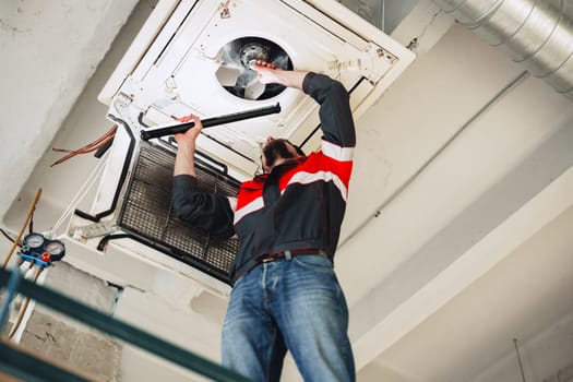 A technician is inspecting and repairing an air conditioning unit on the ceiling of a commercial building in the afternoon. Tools are visible, and the technician shows focus and concentration.