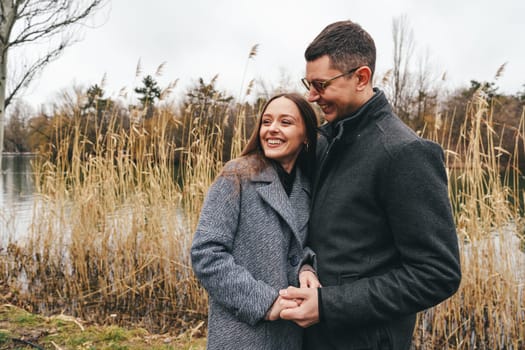 Two people share a joyful embrace near a tranquil lake, surrounded by tall grasses on a cool autumn day. Their smiles reflect happiness and warmth amidst the overcast sky.