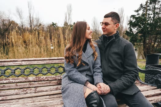A couple sits closely on a bench in a park, sharing smiles as they enjoy a chilly afternoon in late fall surrounded by tall grass and trees.