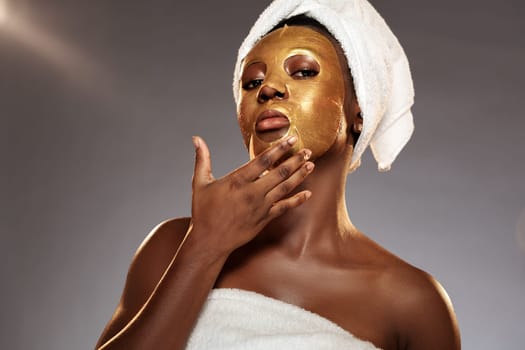 A woman is engaged in a skincare routine, applying a gold facial mask to her face. She is wearing a towel on her head and a white wrap. The lighting creates a warm ambiance.