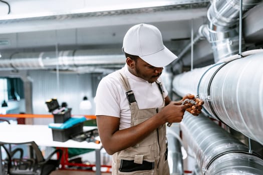 A young technician wearing a cap and overalls works diligently on repairing a ventilation system in a bright, contemporary workshop. Tools and equipment are visible in the background.
