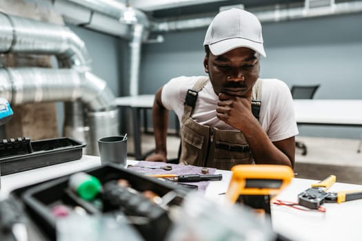 A young man is focused on a collection of tools and equipment laid out on a table in a workshop. He appears to be thinking critically about the task at hand.