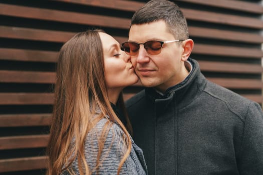 A man and woman embrace affectionately outdoors, with the woman giving a light kiss to the man's cheek. They are standing closely, dressed casually, enjoying the moment.